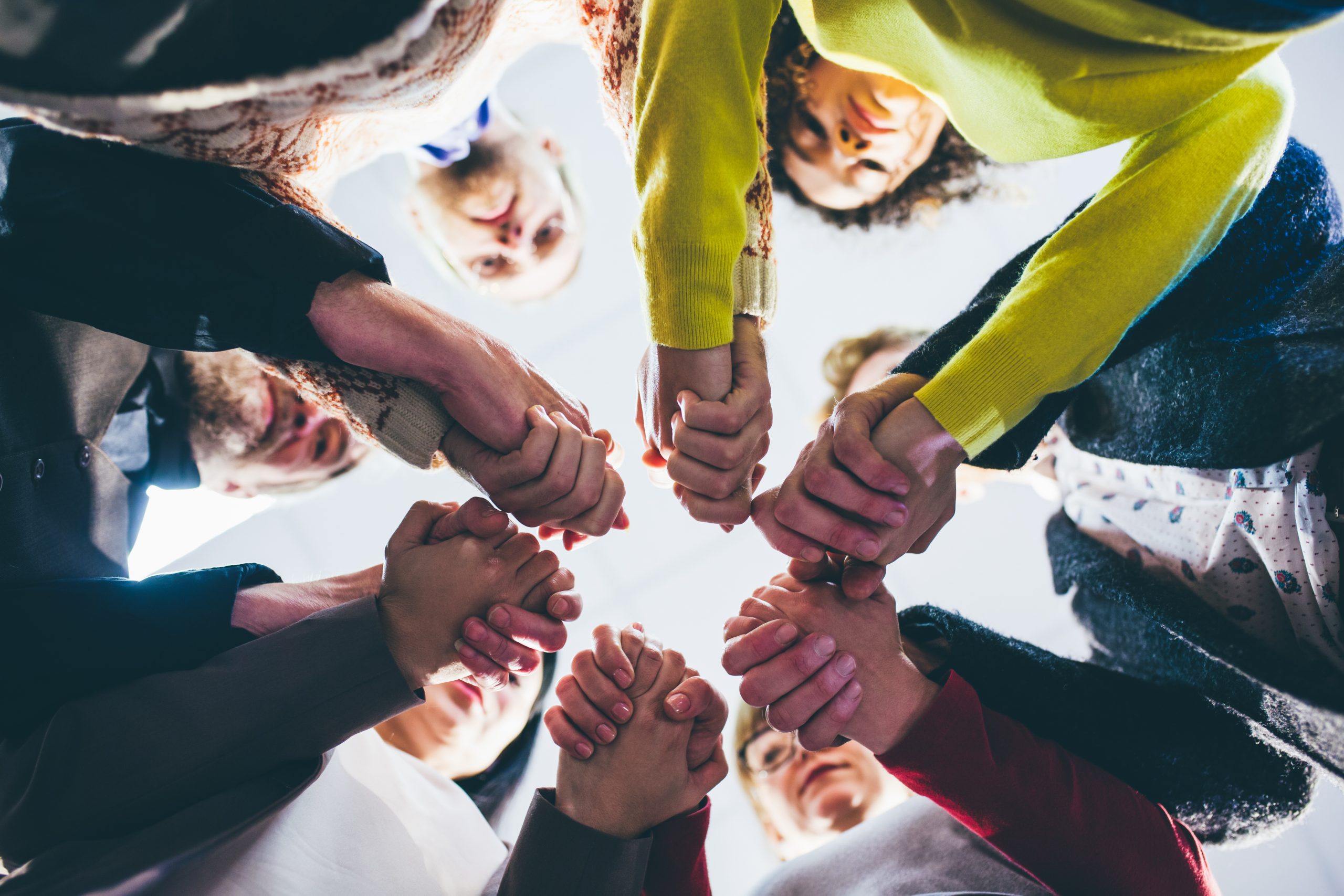Viewed from below, a circle of people of different ages join hands together against a bright sky.