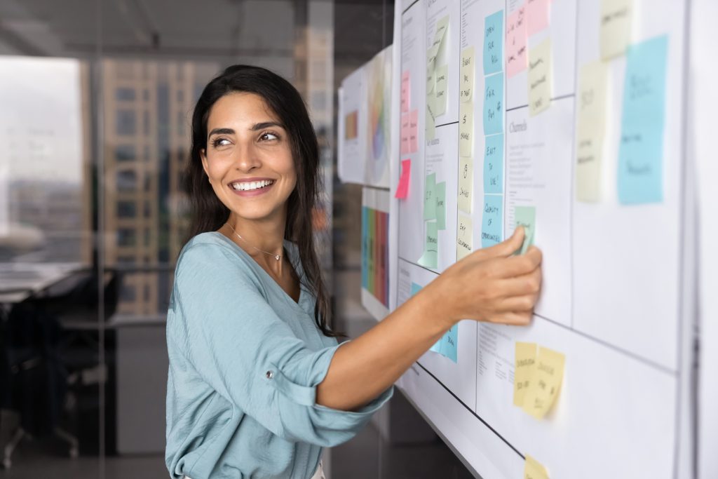 A woman smiles as she places a note on a wall-mounted planning board covered in colored cards.