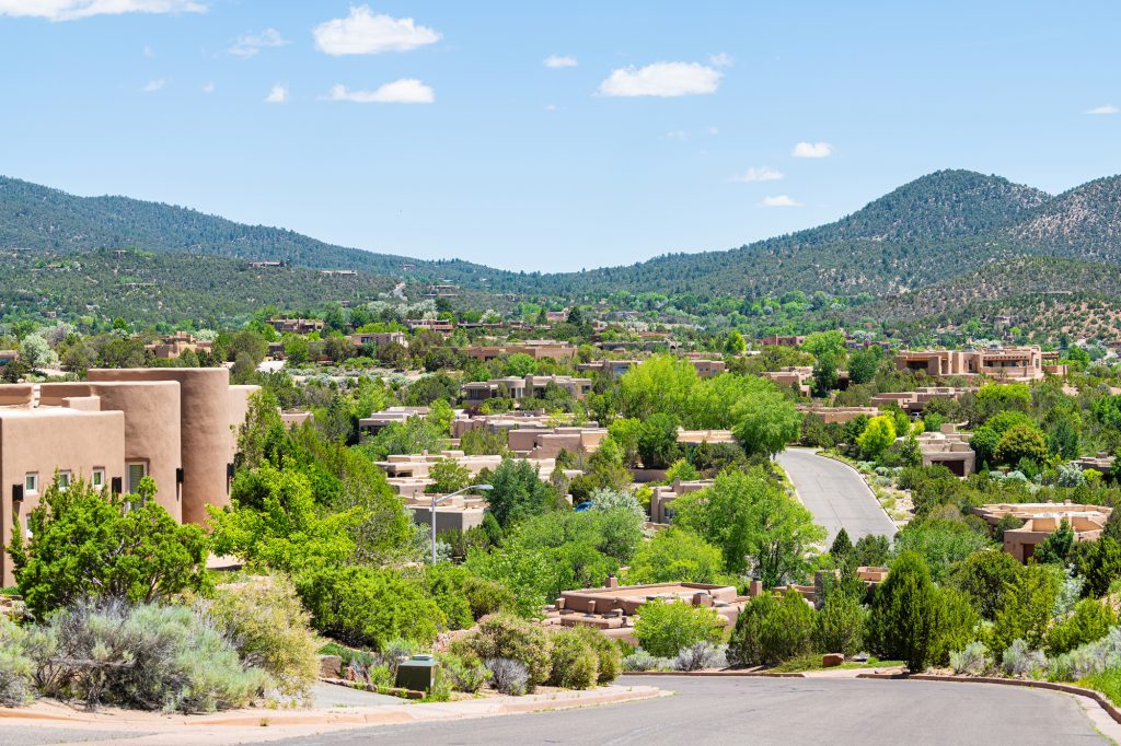 A view across Santa Fe of low adobe homes among green pinon and juniper, with blue mountains behind.