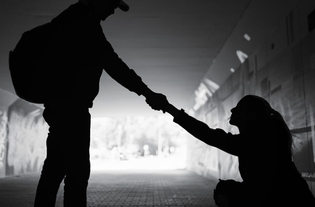 In silhouette, a person carrying a backpack reaches down to help someone up off the ground.