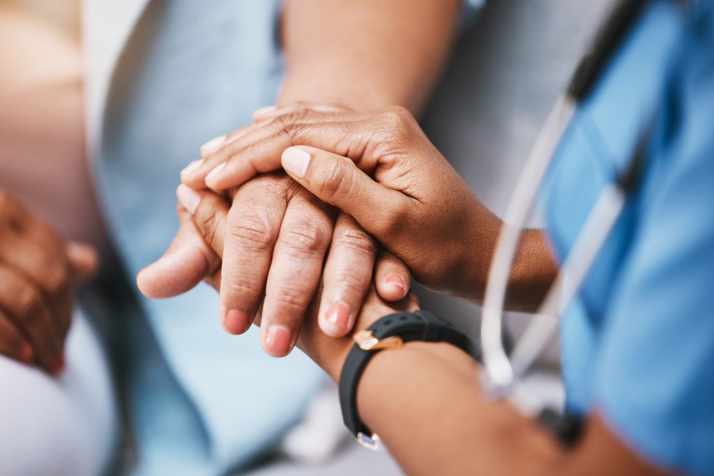 A caregiver in scrubs holds an older person's hands in both of hers.