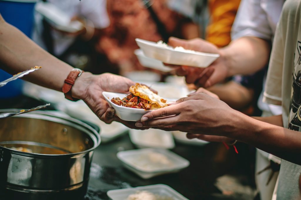 Hands pass a bowl of hot food across a serving table lined with pots and empty bowls.